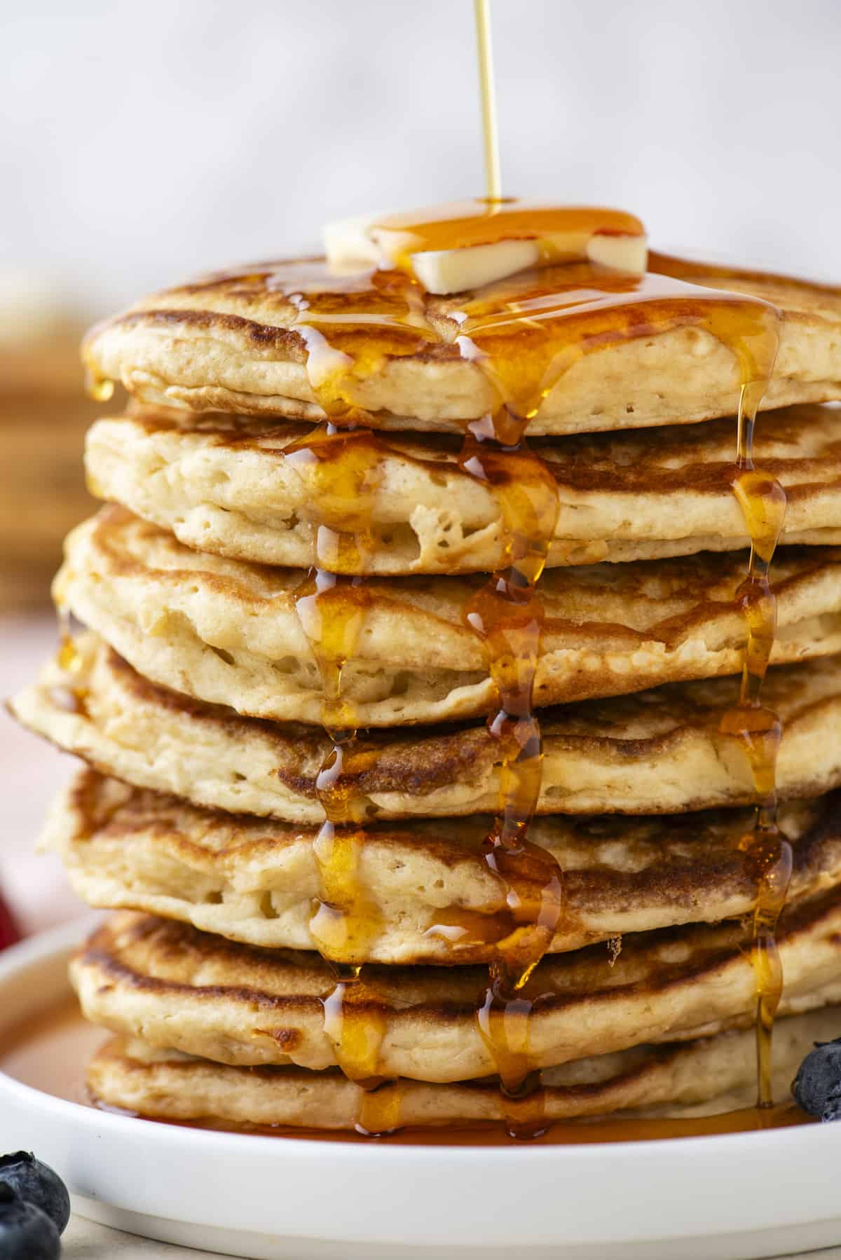 a tall stack of pancakes on a white plate being drizzled with syrup, topped with a slice of butter and surrounded by fresh berries