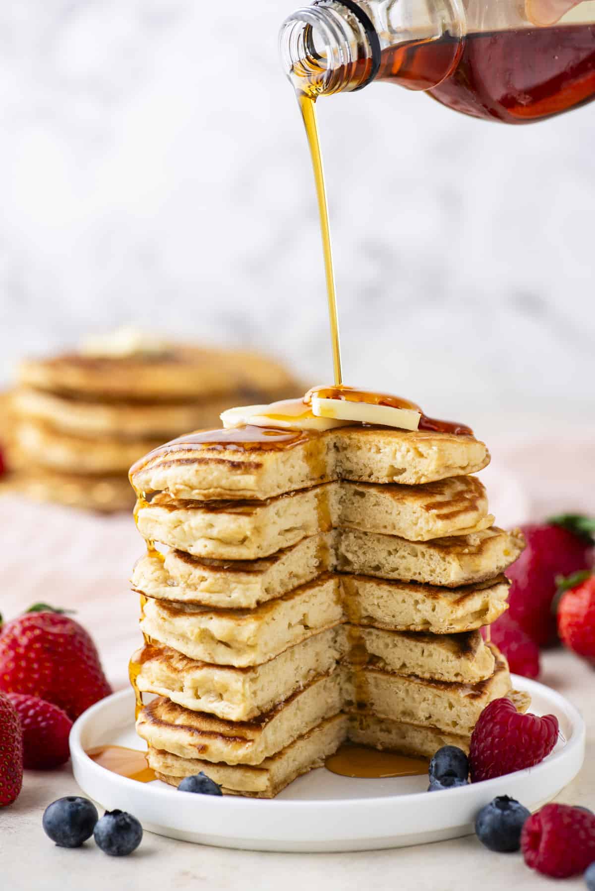 a tall stack of pancakes with a triangular portion cut out of the entire stack, being drizzled with syrup, topped with a slice of butter and surrounded by fresh berries on a white plate