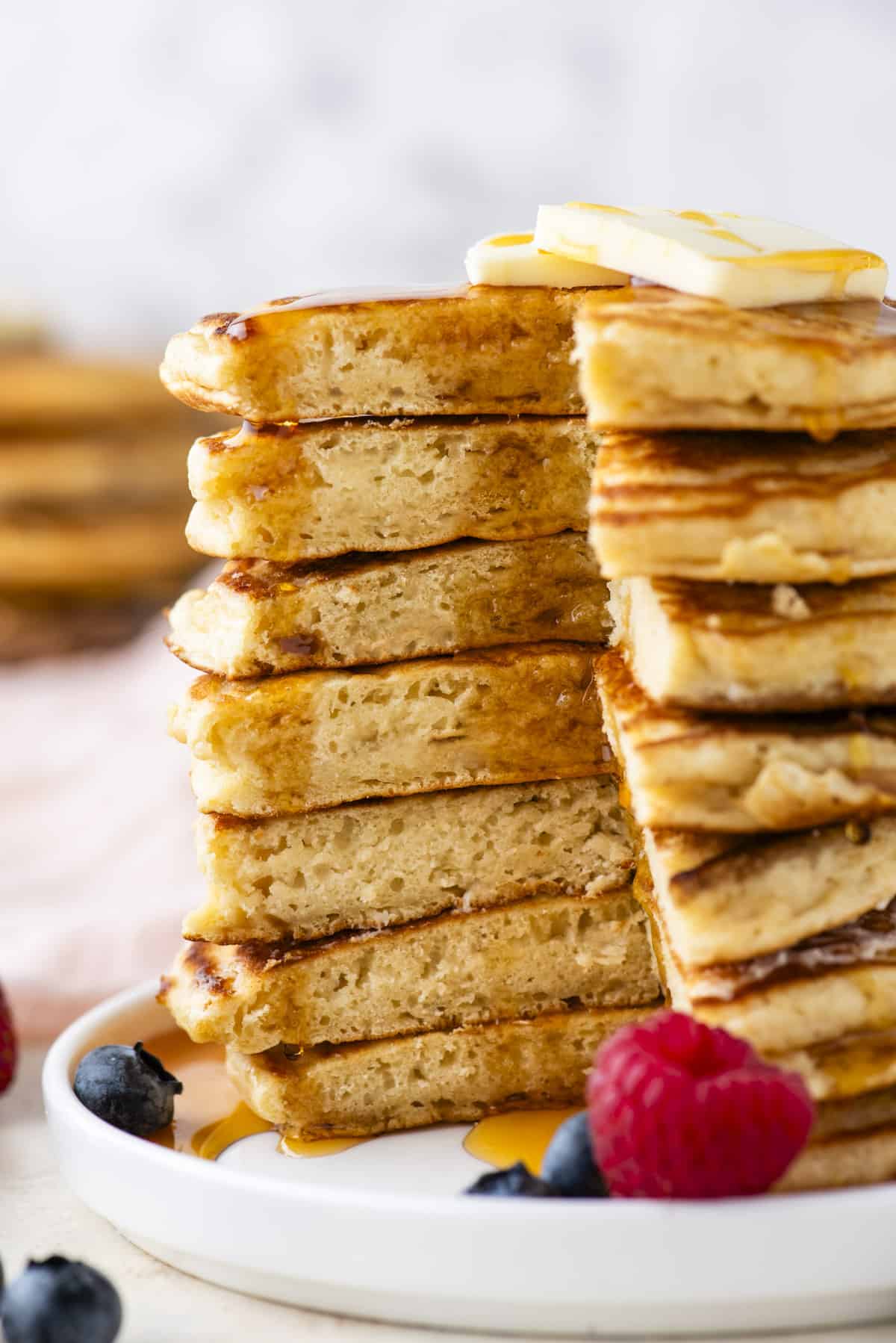 a tall stack of pancakes with a triangular portion cut out of the entire stack, drizzled with syrup, topped with a slice of butter and surrounded by fresh berries on a white plate