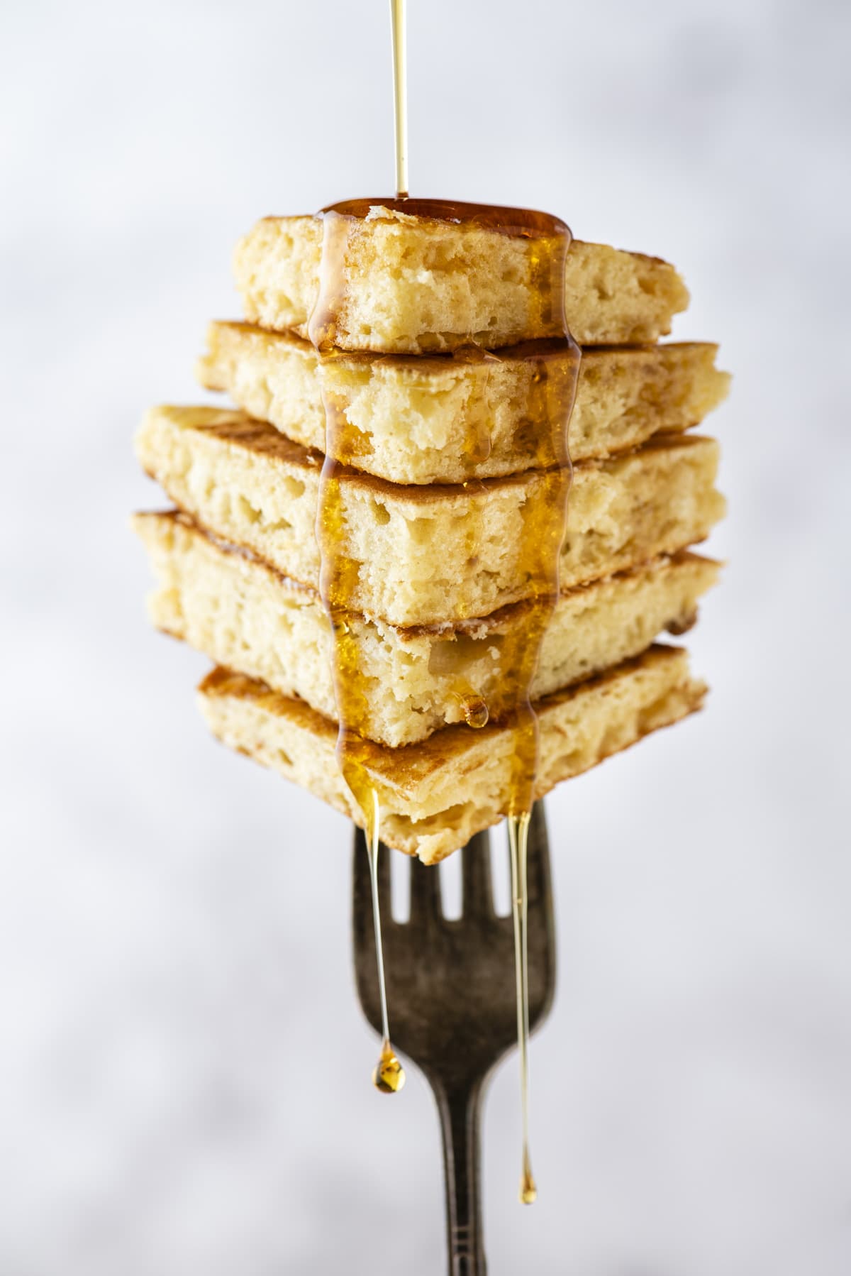 a stack of pancake slices piled up and being held vertically on a fork, with syrup being drizzled and dripping down