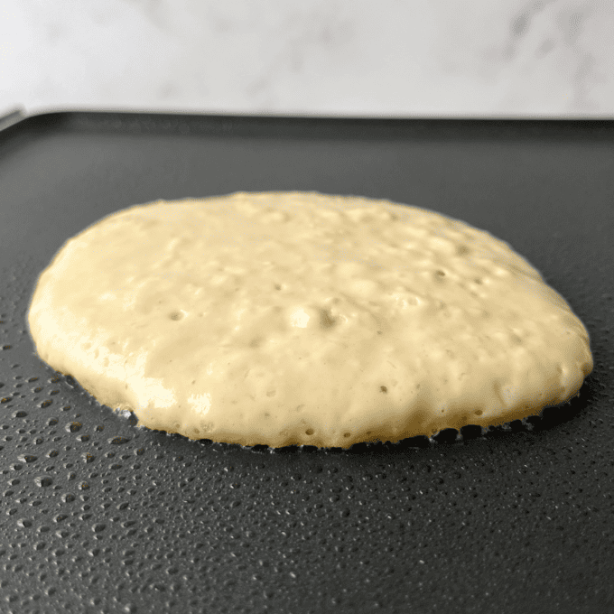 a pancake cooking on a griddle that hasn't been flipped yet, with bubbles starting to form on the top
