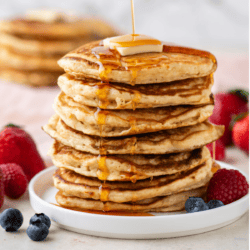 a tall stack of pancakes on a white plate being drizzled with syrup, topped with a slice of butter and surrounded by fresh berries, with more pancakes in the background