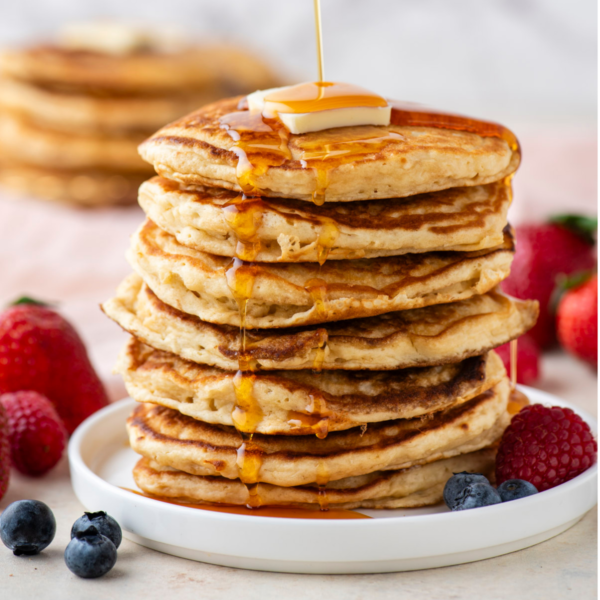 a tall stack of pancakes on a white plate being drizzled with syrup, topped with a slice of butter and surrounded by fresh berries, with more pancakes in the background