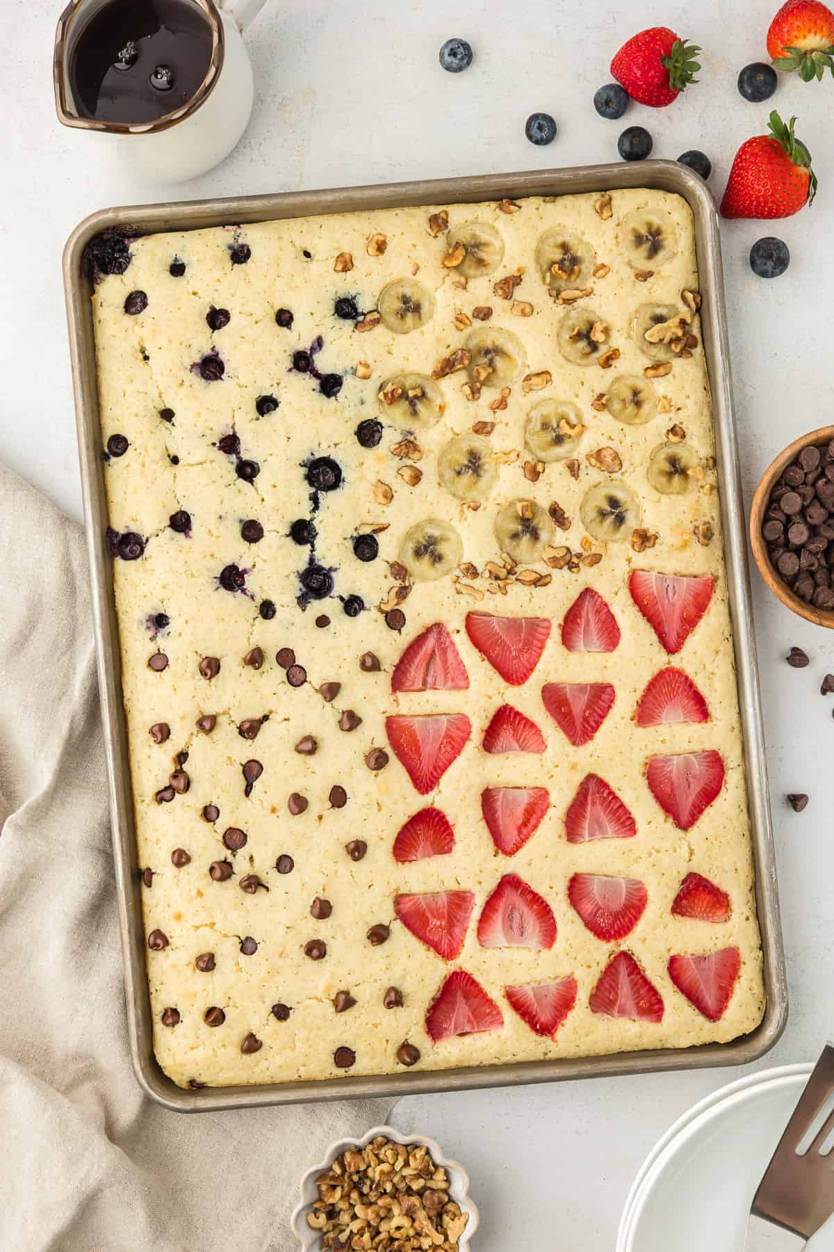 A sheet pan full of pancakes, blueberry pancakes in the upper left corner, banana walnut pancakes in the upper right corner, chocolate chip pancakes in the lower left corner and strawberry pancakes in the lower right corner, with syrup, fresh berries, a cup of chocolate chips, a cup of chopped nuts and a light tan towel surrounding the sheet pan
