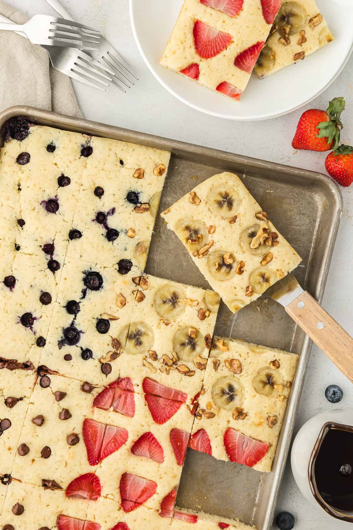 A sheet pan full of pancakes, some with blueberries in them, some with bananas and nuts, some with chocolate chips and some with strawberries, with one pancake being lifted by a spatula, syrup, fresh blueberries and strawberries, forks, naples, and a plate with two slices of pancake around the sheet pan