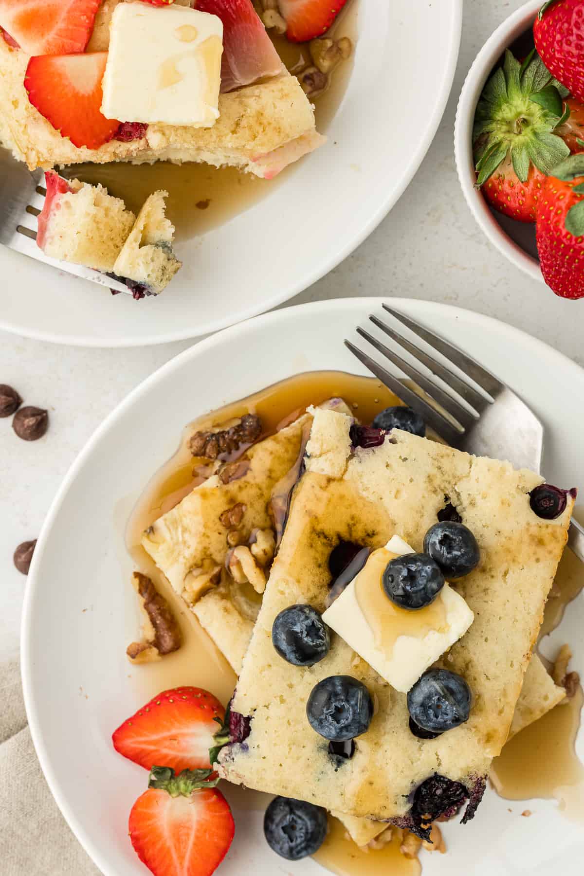 two white plates with slices of sheet pan pancakes on them, topped with slices of butter, maple syrup and fresh blueberries and strawberries, with forks on both plates and a bowl of fresh stawberries beside them