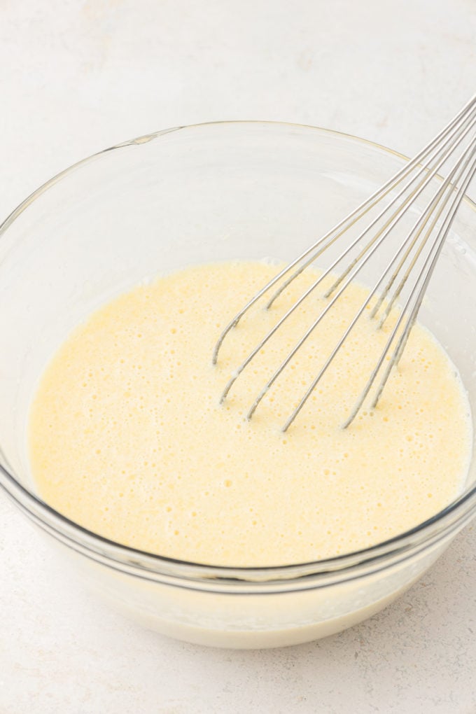 wet ingredients for sheet pan pancakes in a clear glass bowl with a whisk leaning inside the bowl