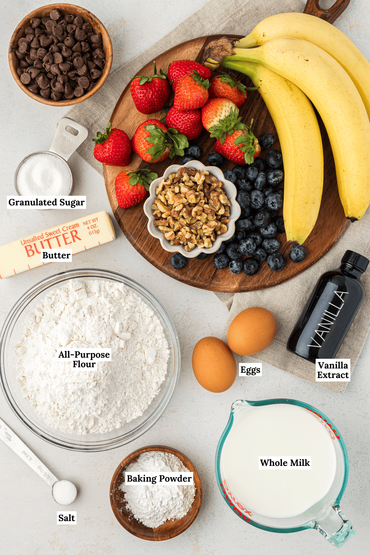 over-head view of ingredients for sheet pan pancakes including whole milk, baking powder, vanilla extract, salt, eggs, all-purpose flour, butter, granulated sugar, chocolate chips, bananas, strawberries, blueberries and nuts