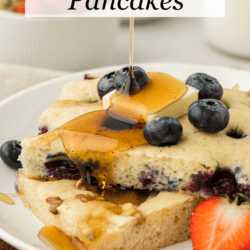 two slices of sheet pan pancakes on a white plate topped with fresh blueberries, a slice of butter and being drizzled with syrup, with a fresh strawberry slice on the plate beside the pancakes