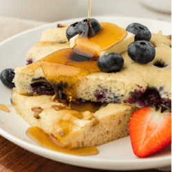 two slices of sheet pan pancakes on a white plate topped with fresh blueberries, a slice of butter and being drizzled with syrup, with a fresh strawberry slice on the plate beside the pancakes