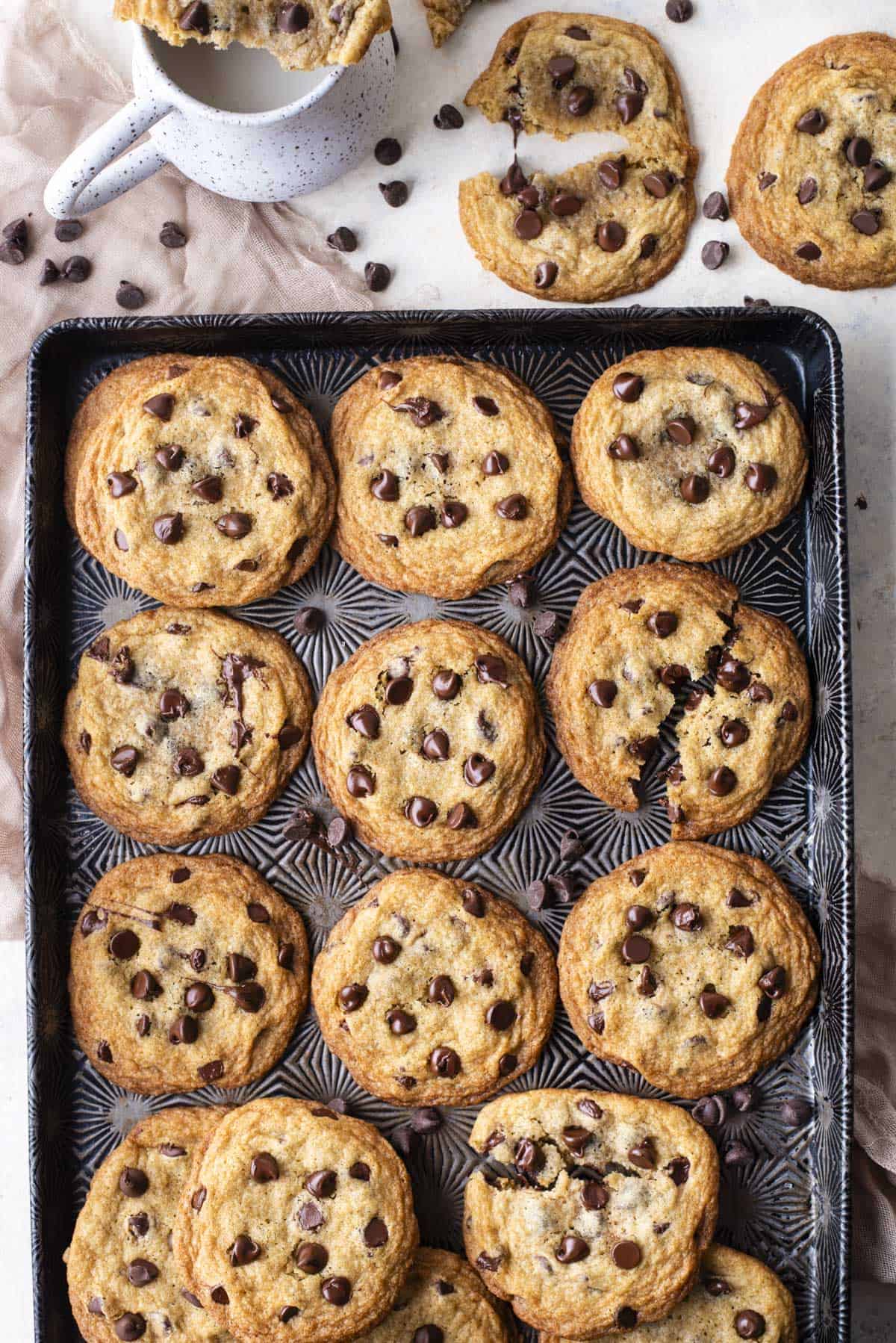 chocolate chip cookies in rows filling a dark metal sheet pan on top of a pink cloth, surrounded by chocolate chips sprinkled around, a whole cookie and a cookie that is partially pulled in half, and a piece of a cookie resting on top of a white mug of milk