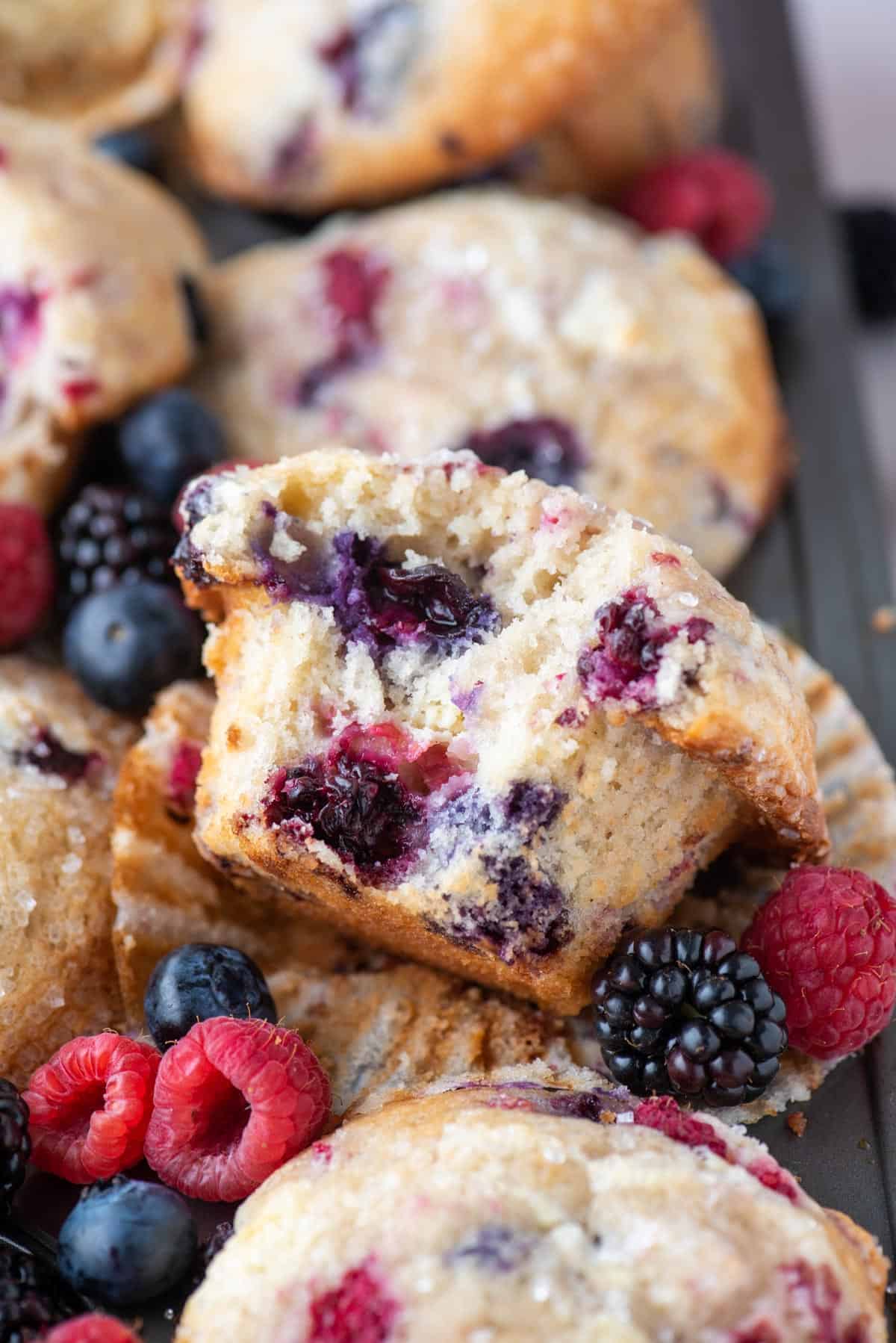a triple berry cheesecake muffin missing one bite that exposes it's center, laying on its open muffin liner with raspberries, blackberries and blueberries around it, on top of a muffin pan full of more muffins
