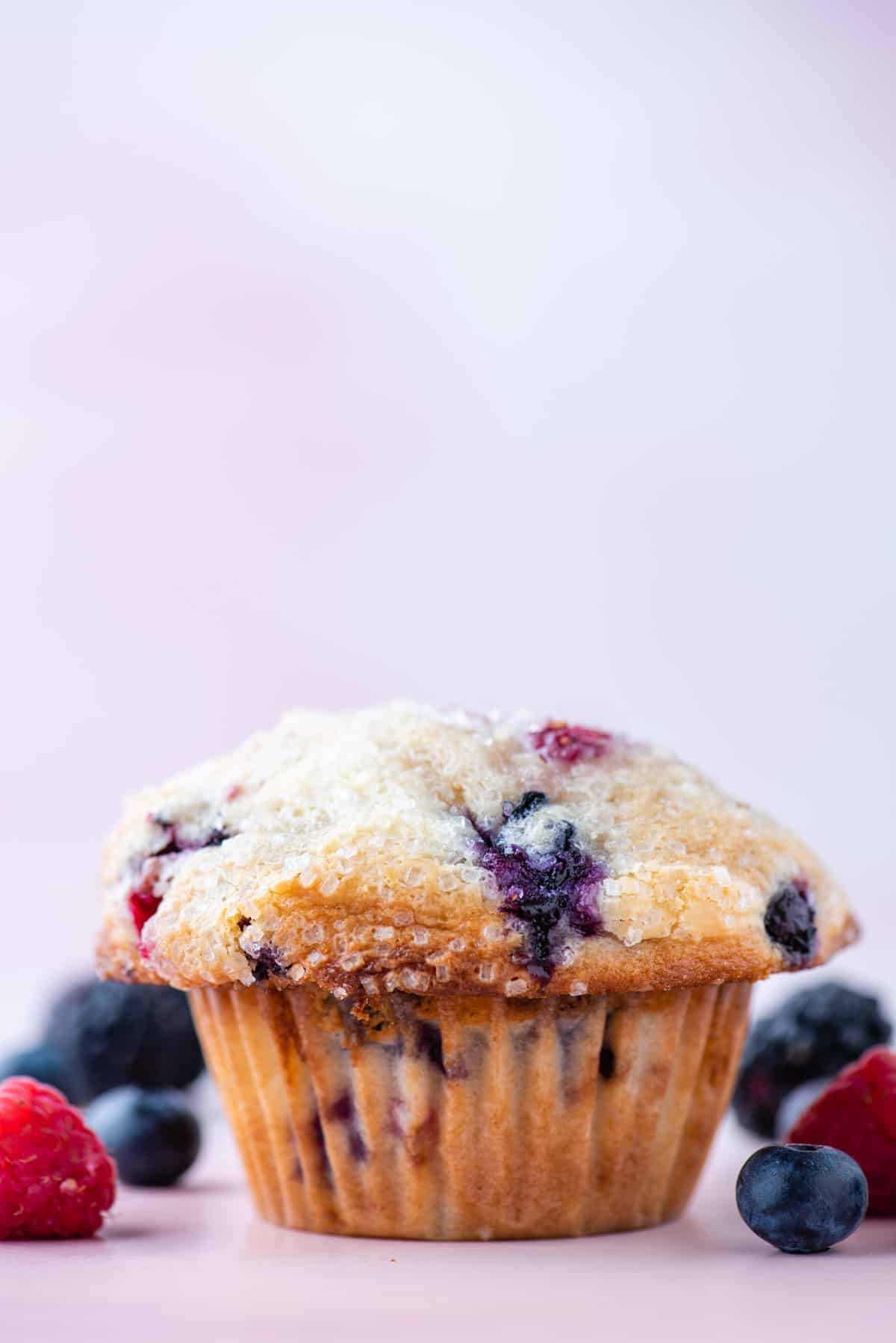 a triple berry cheesecake muffin sitting on a light pink surface surrounded by fresh blueberries, blackberries and raspberries