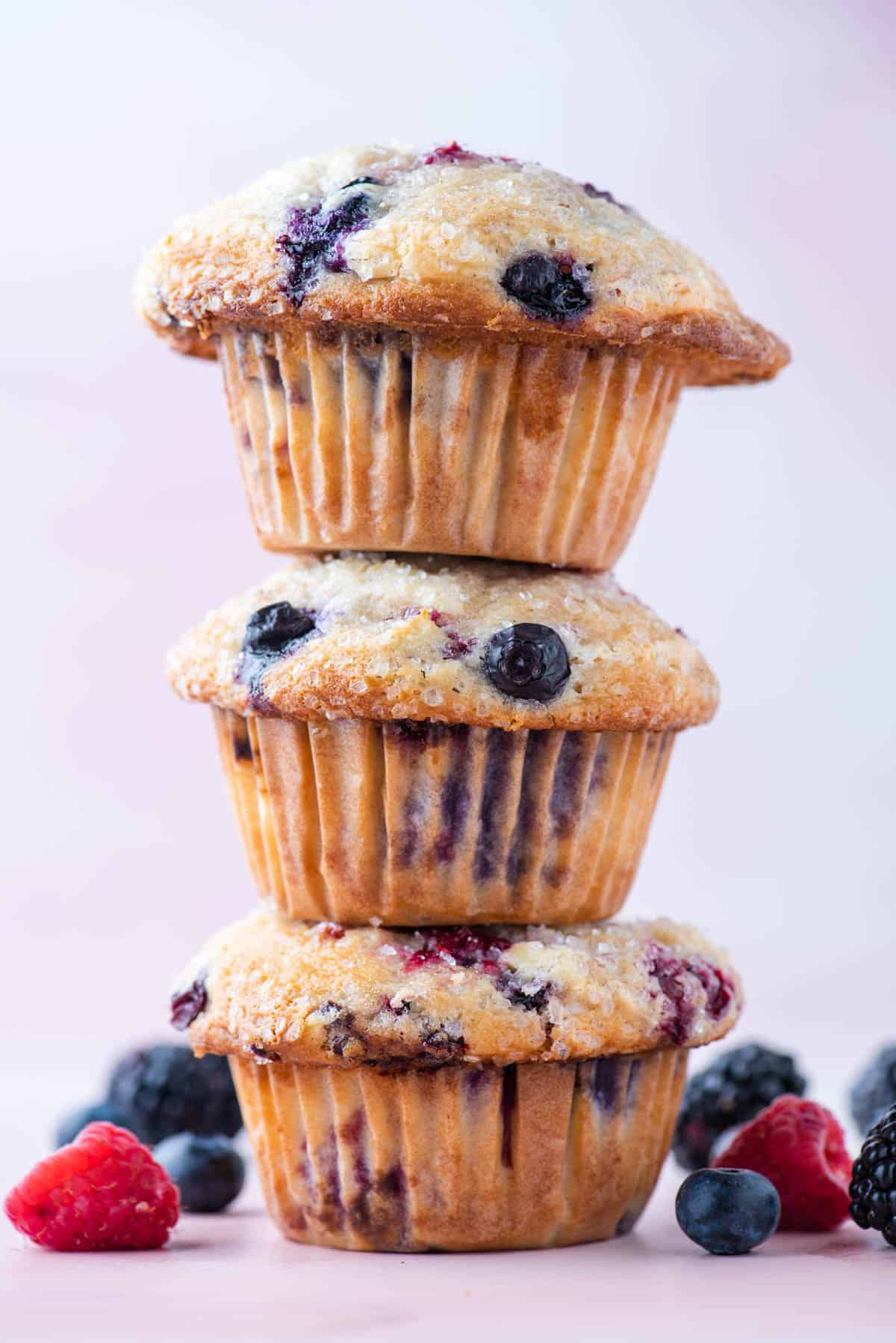 a stack of 3 triple berry cheesecake muffins sitting on a pink surface, surrounded by fresh blueberries, blackberries and raspberries