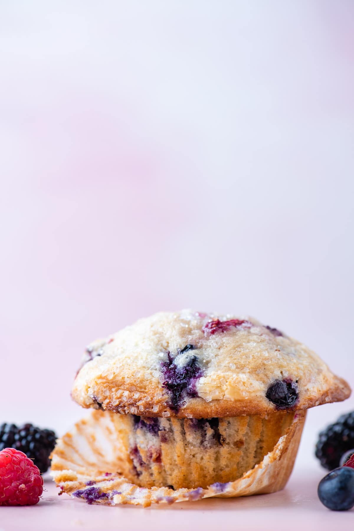 a triple berry cheesecake muffin sitting on a light pink surface with its muffin liner partially peeled away, surrounded by fresh blueberries, blackberries and raspberries