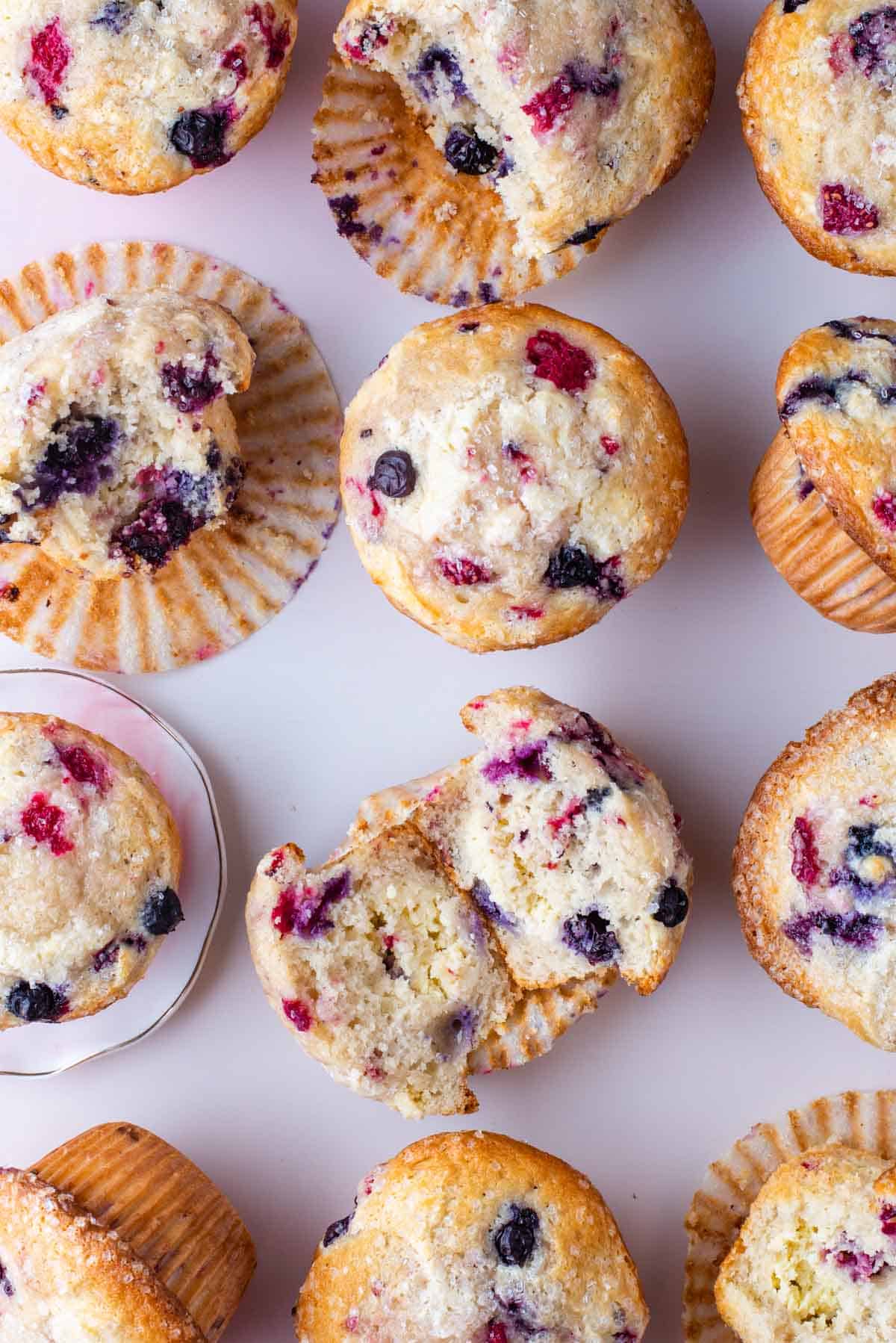 rows of triple berry cheesecake muffins on a light pink surface, some sitting upright, some on their sides, some cut in half and some with a bite out