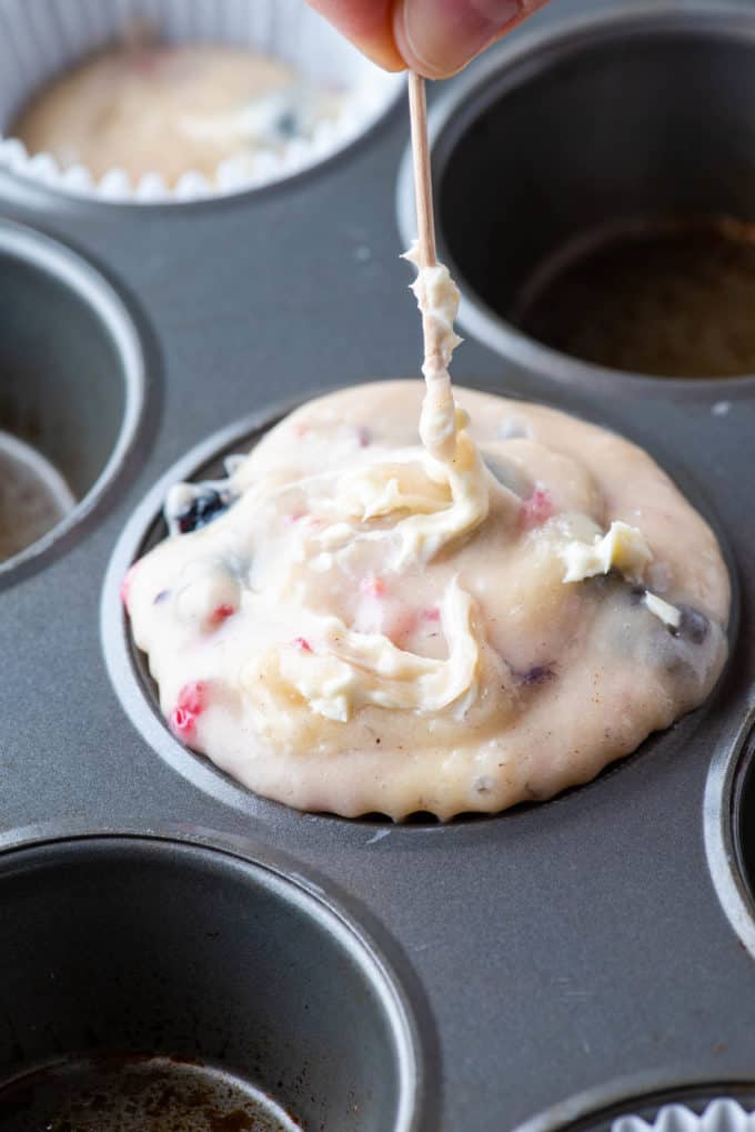 triple berry cheesecake muffin batter in a muffin liner in a muffin pan with cream cheese filling being swirled into the top with a toothpick