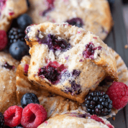 a triple berry cheesecake muffin missing one bite that exposes it's center, laying on its open muffin liner with raspberries, blackberries and blueberries around it, on top of a muffin pan full of more muffins
