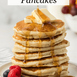 a tall stack of buttermilk pancakes on a white plate with two slices of butter on top and fresh blueberries and raspberries on the plate being drizzled with maple syrup