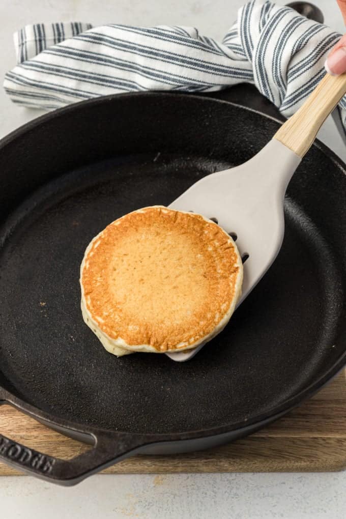a buttermilk pancake being flipped with a spatula over a cast iron skillet that's held by the handle with a blue and white striped towel
