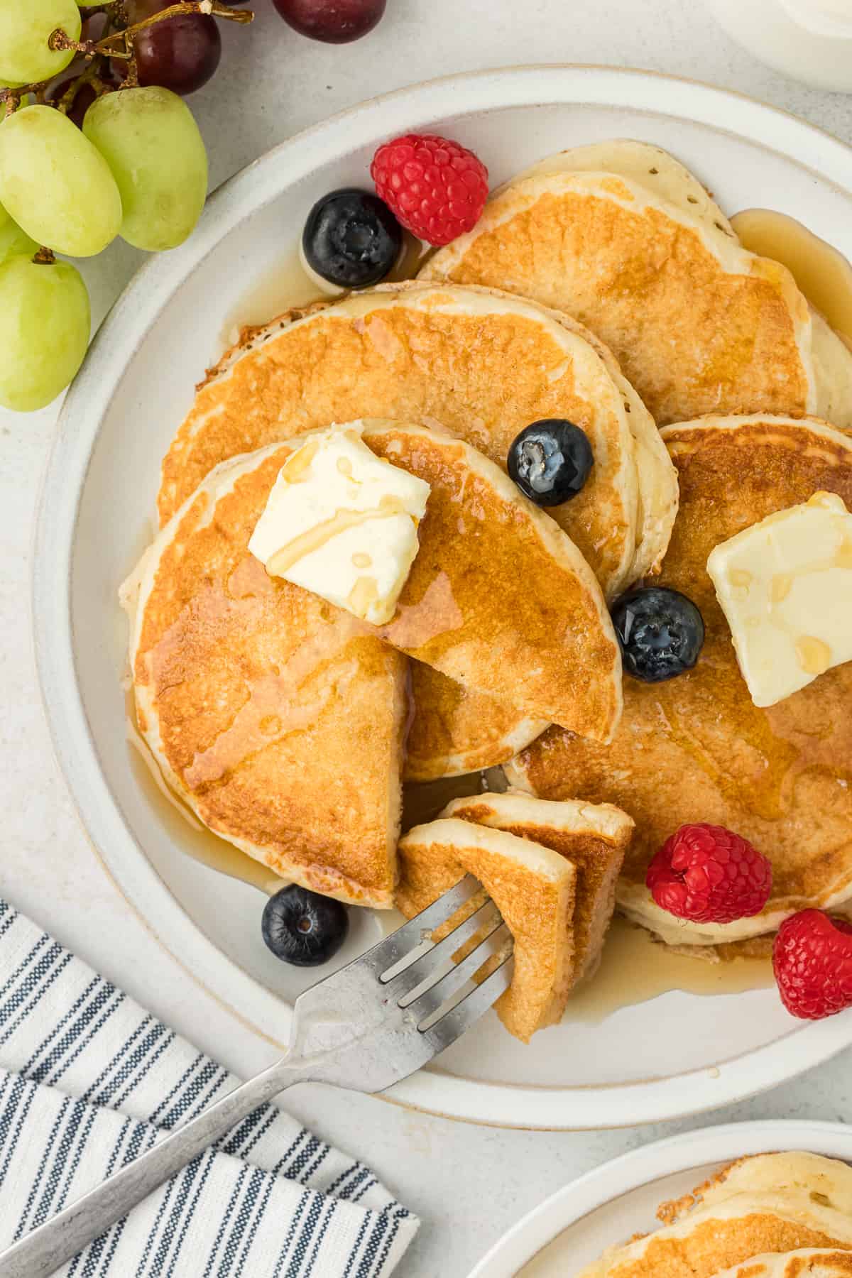 a white plate with four buttermilk pancakes on it, with a triangle-shaped slice out of two pancakes on the tip of a fork that's resting on the plate, fresh blueberries and raspberries, butter slices and drizzles of syrup topping the pancakes, a blue and white striped towel, fresh grapes and another plate of pancakes beside the plate of pancakes