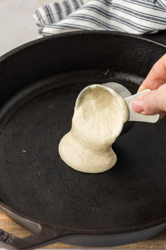 Buttermilk pancake batter being poured onto a cast iron skillet with a blue and white striped towel beside it