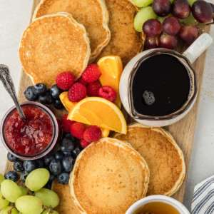 a wooden cutting board covered in buttermilk pancakes, a bowl of serving, a small bowl of jam with a spoon in it, a small bowl of honey, and fresh orange slices, raspberries, blueberries and green and red grapes