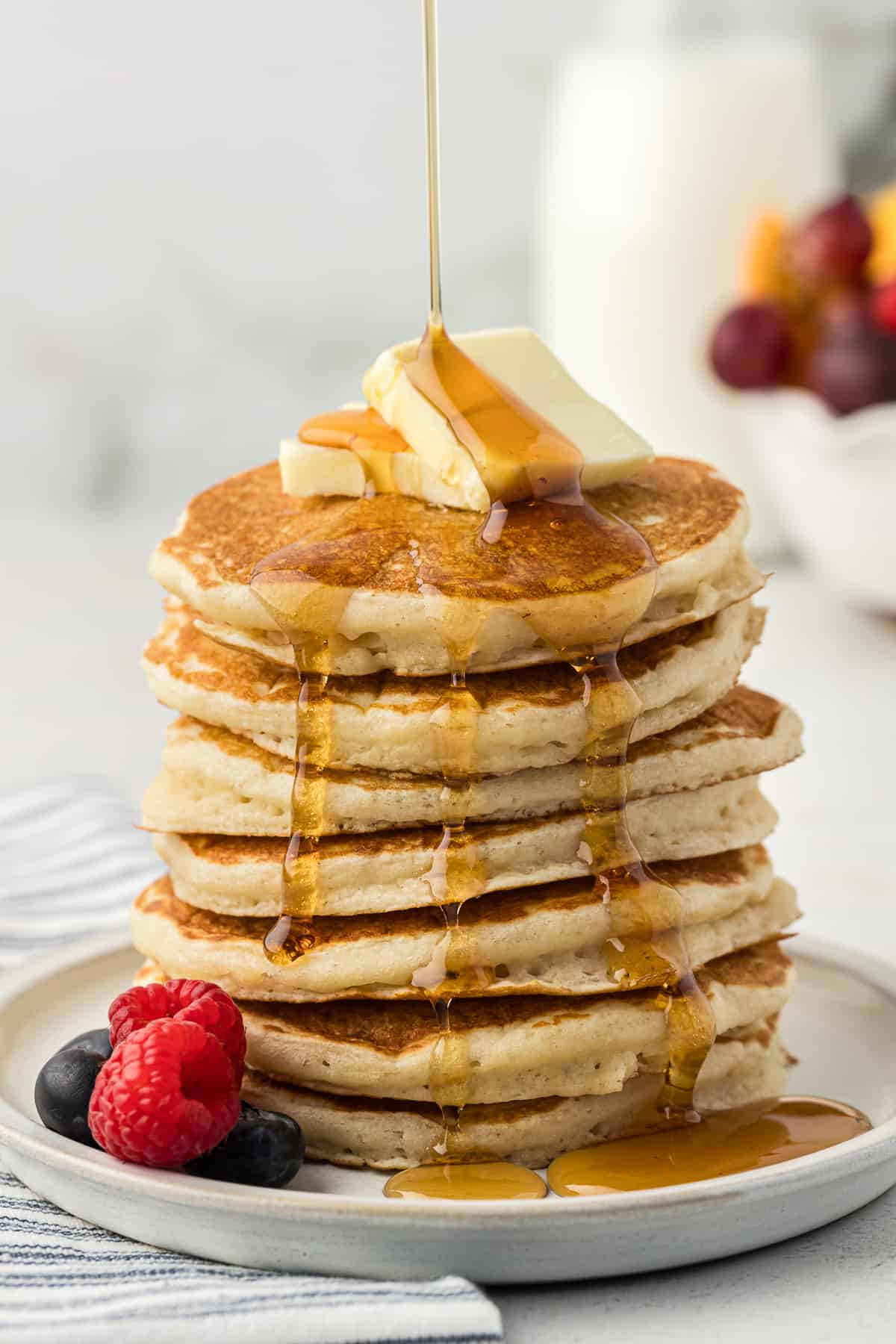 a tall stack of buttermilk pancakes on a white plate with two slices of butter on top and fresh blueberries and raspberries on the plate being drizzled with maple syrup