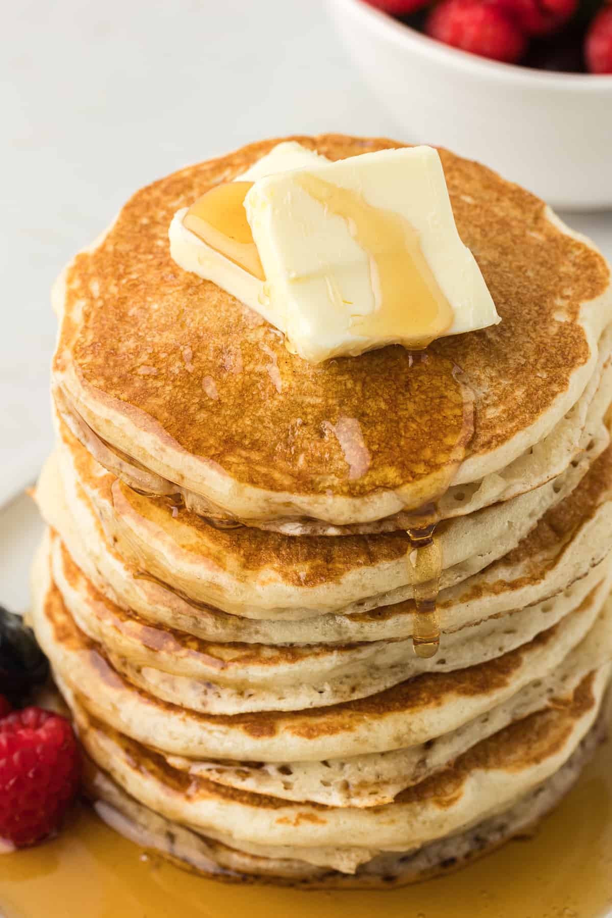 a tall stack of buttermilk pancakes on a white plate with two slices of butter on top and fresh blueberries and raspberries on the plate drizzled with maple syrup