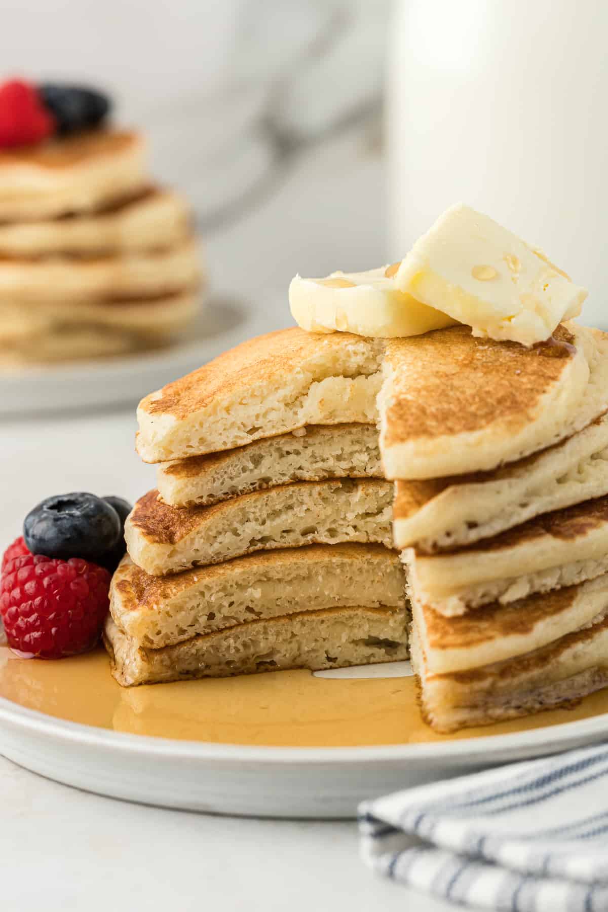 a white plate with 5 pancakes in a stack, with one large slice out of the entire stack, fresh blueberries and raspberries beside the pancakes, two slices of butter on top and syrup all over the plate