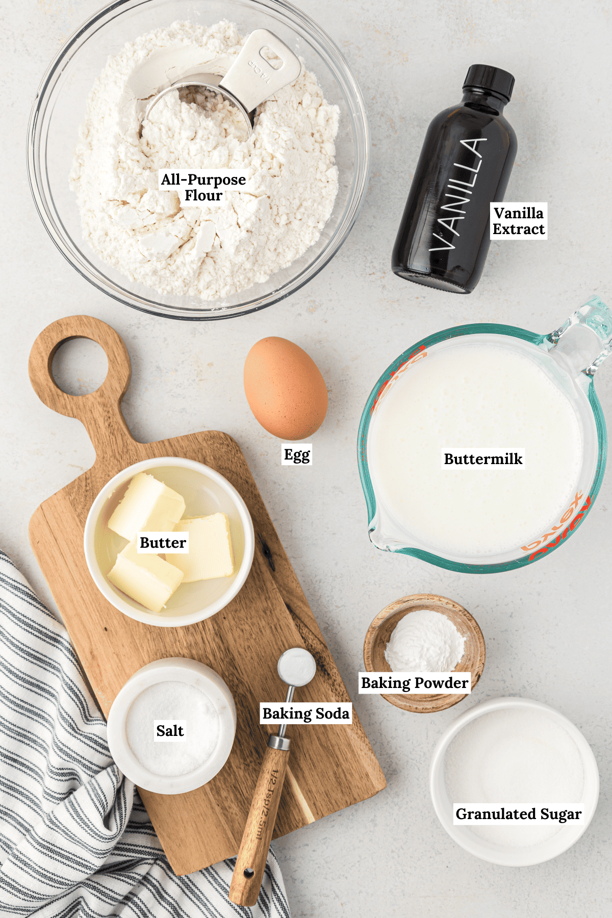 overhead view of the ingredients for buttermilk pancakes including all-purpose flour, vanilla extract, one large egg, buttermilk, baking powder, butter, salt, baking soda and granulated sugar