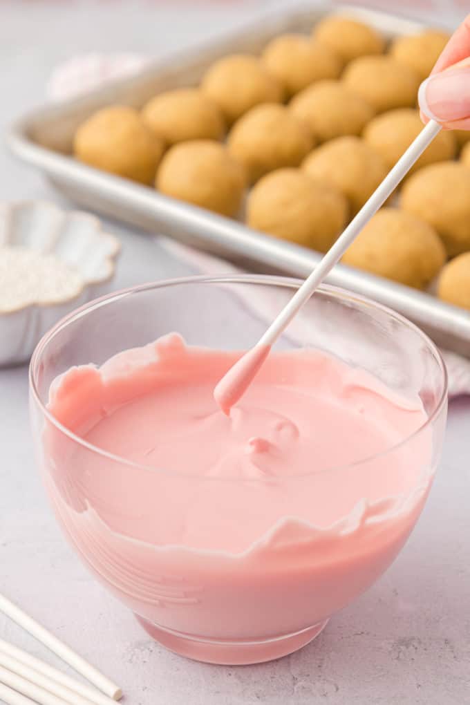 a cake pop stick that has just been dipped in pink candy melt over top of a clear glass bowl of melted candy melts, with a sheet pan of plain cake pops behind it