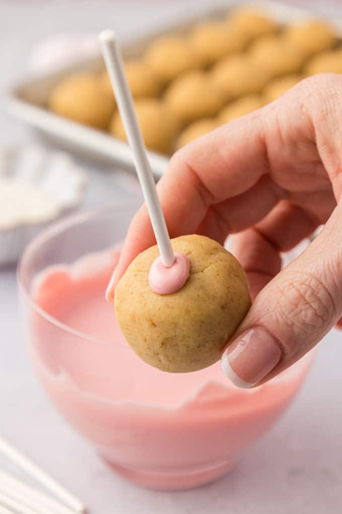 a hand holding a cake pop on a cake pop stick with a bit of pink candy melts around where the stick entered the cake pop, with a bowl of pink candy melts and a sheet pan of plain cake pops in the background