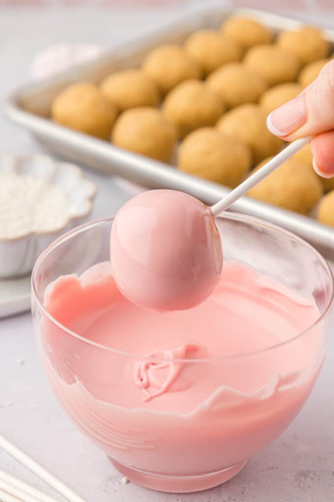 a freshly dipped pink cake pop over top of a clear glass bowl full of melted pink candy melts with a sheet pan of plain cake balls in the background