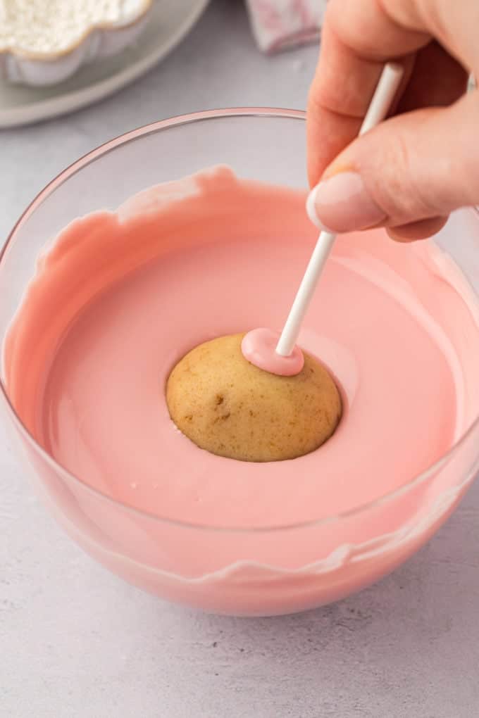 a cake pop on a white stick being dipped into a clear glass bowl full of melted pink candy melts