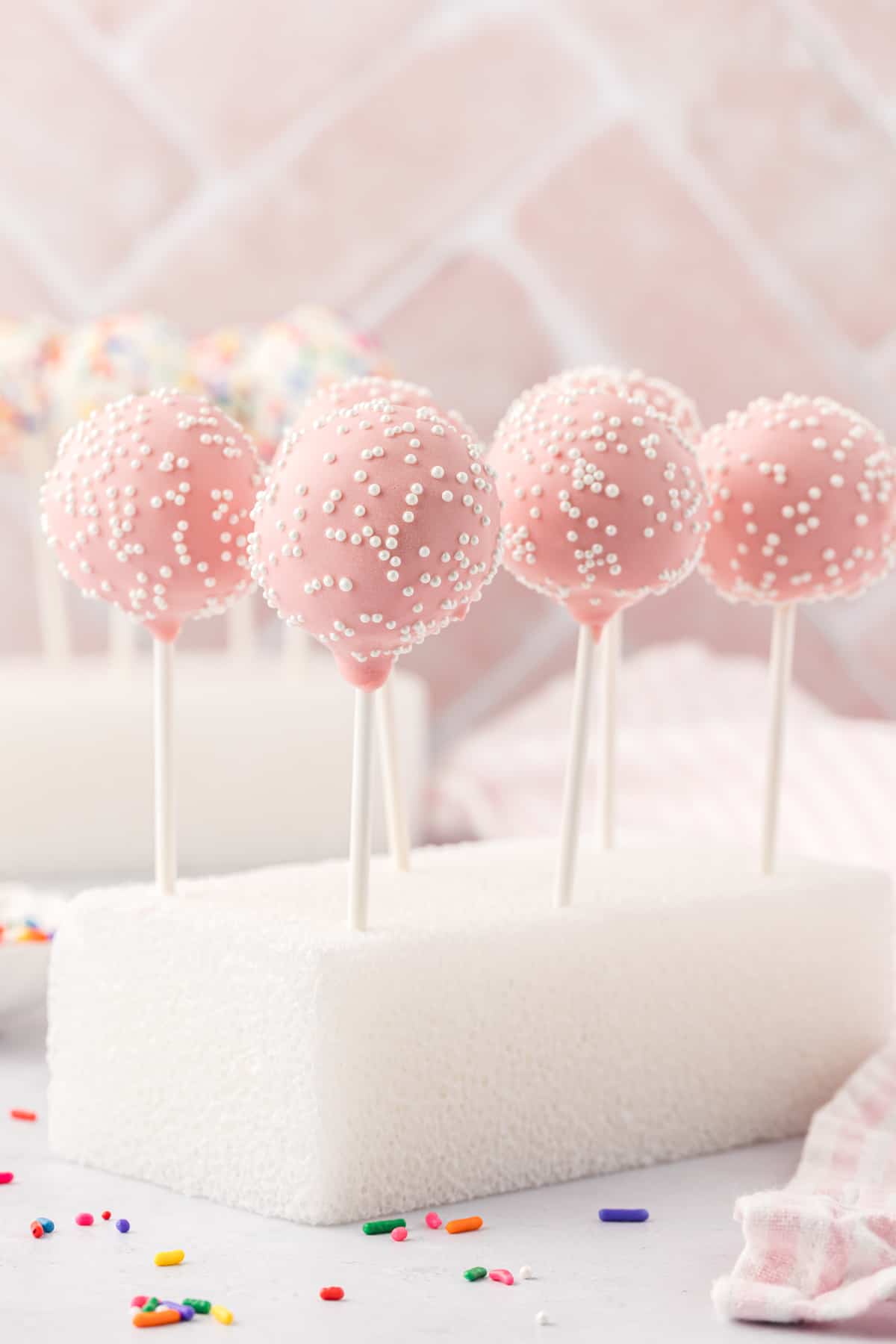 a styrofoam block with 6 pink cake pops with white sprinkles stuck in it so they stand straight up, with a white and pink striped towel and rainbow sprinkles scattered around it