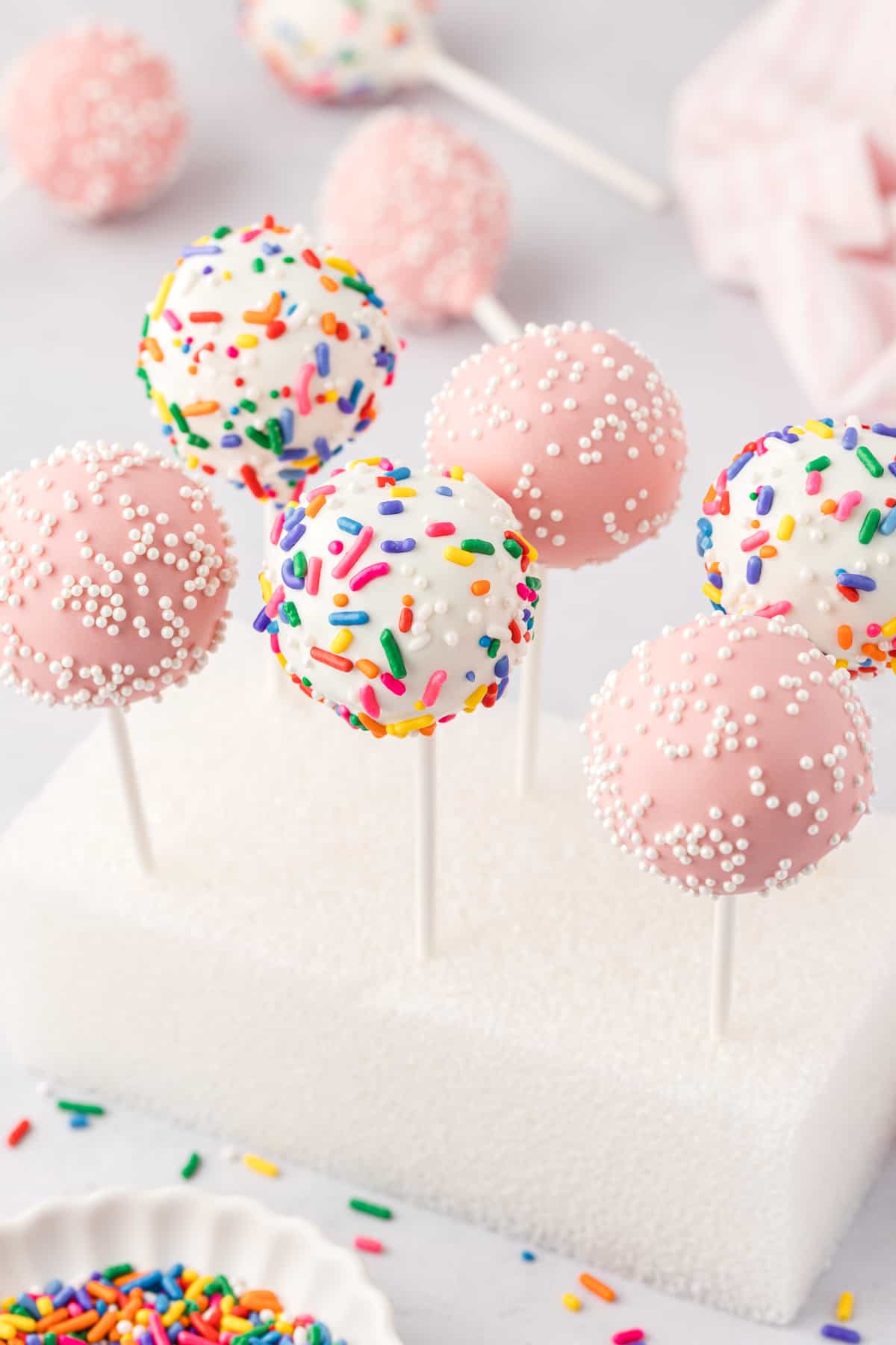 a styrofoam block with six cake pops stuck in it so they stand straight up, three pink with white sprinkles and three white with rainbow sprinkles, with a white dish of rainbow sprinkles in the front and more cake pops laying in the background