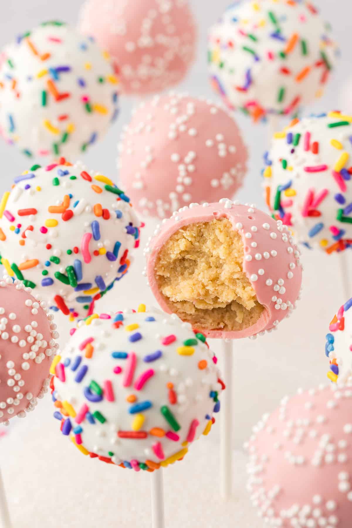 cake pops standing straight up set in a white styrofoam block, some pink with white sprinkles and some white with rainbow sprinkles, with one pink one missing a bite out
