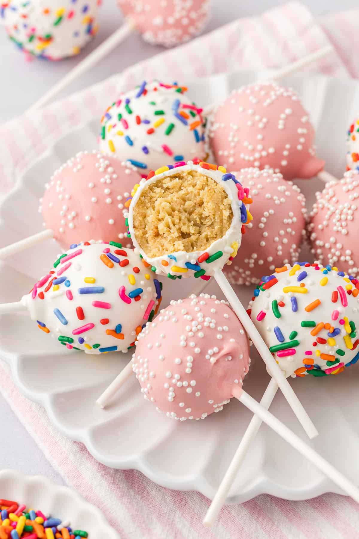 a white plate with a pile of cake pops, some light pink with white sprinkles and some white with rainbow sprinkles, with the cake pop on top missing one bite out of it