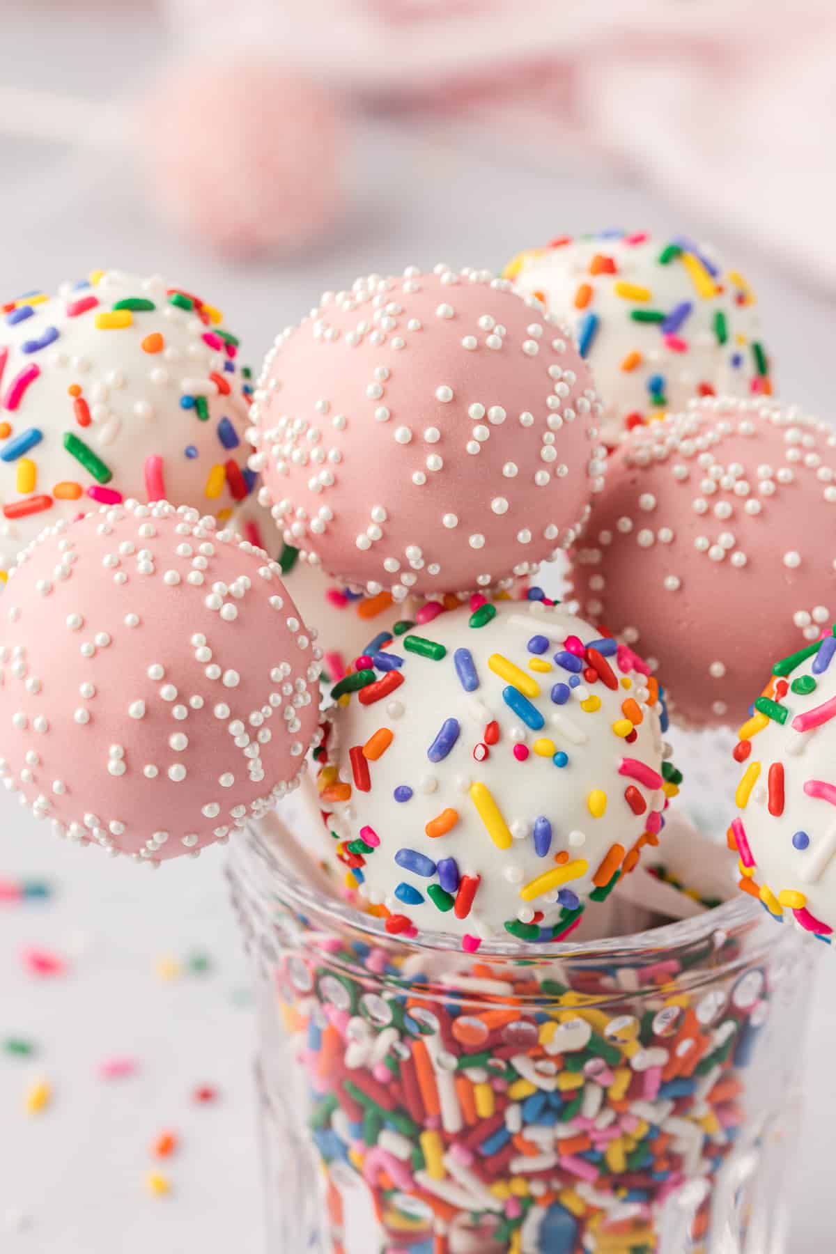a clear glass jar full of rainbow sprinkles with cake pops stuck in the top, some white with rainbow sprinkles and some pink with white sprinkles