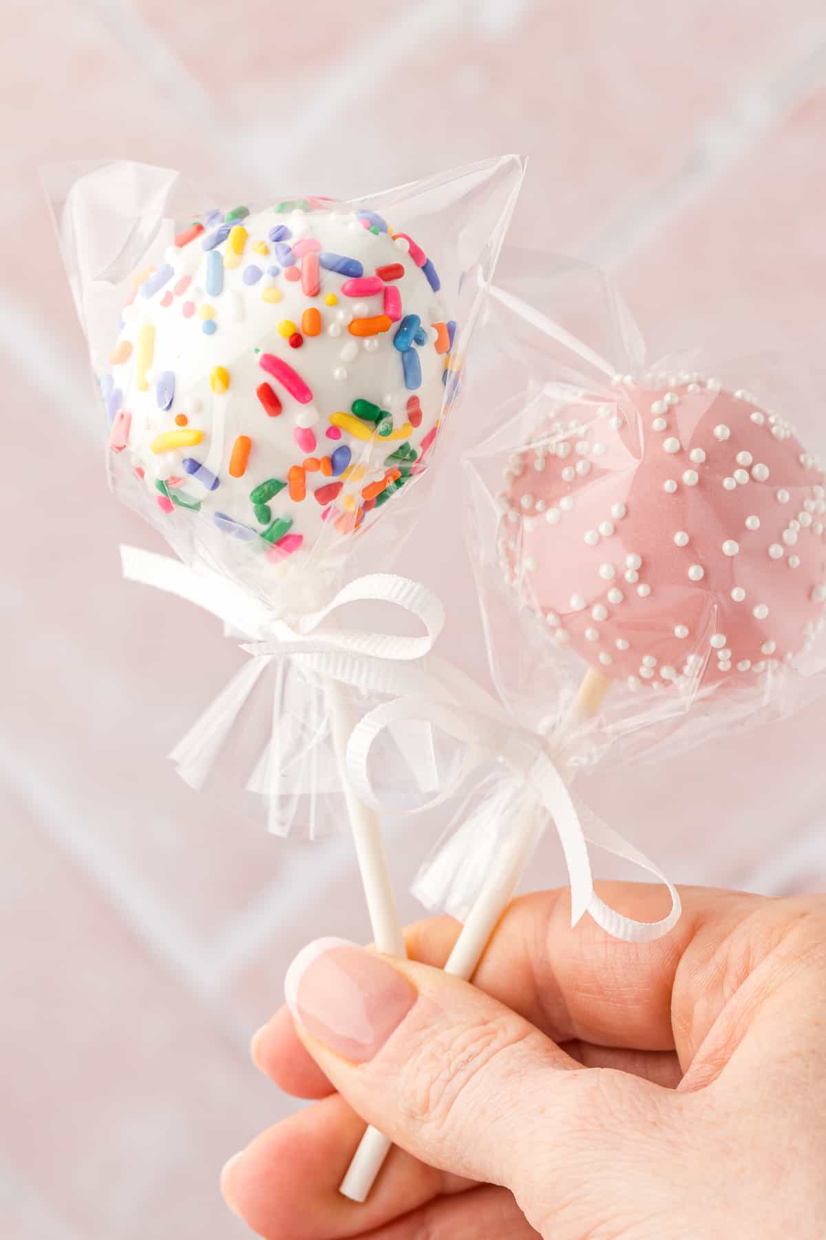 a hand holding two cake pops, one white with rainbow sprinkles and one pink with white sprinkles, both wrapped in plastic and tied with a white ribbon