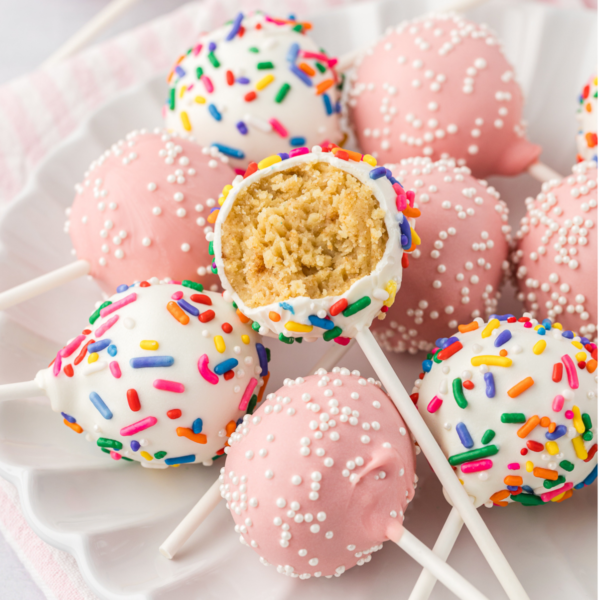 a white plate with a pile of cake pops, some light pink with white sprinkles and some white with rainbow sprinkles, with the cake pop on top missing one bite out of it