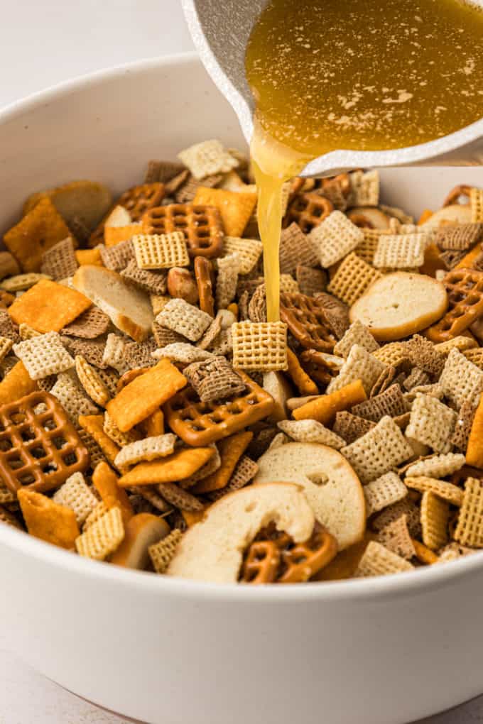 a white bowl full of chex mix ingredients with a seasoned butter mixture being poured in