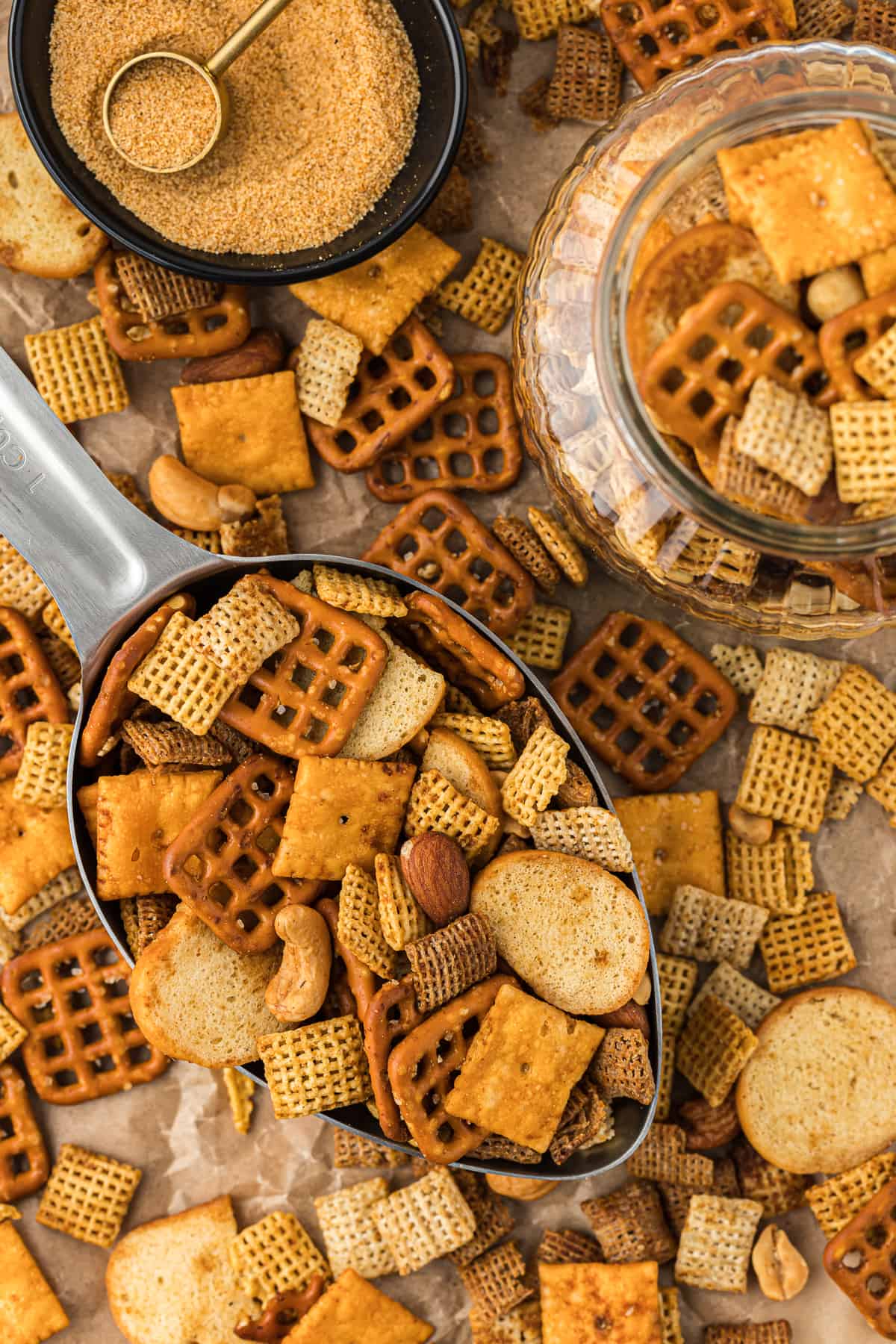 over head view of a large metal scoop full of chex mix on top of a brown paper surface covered in chex mix, a small black bowl of seasoning with a measuring spoon in it, and a clear glass jar of chex mix
