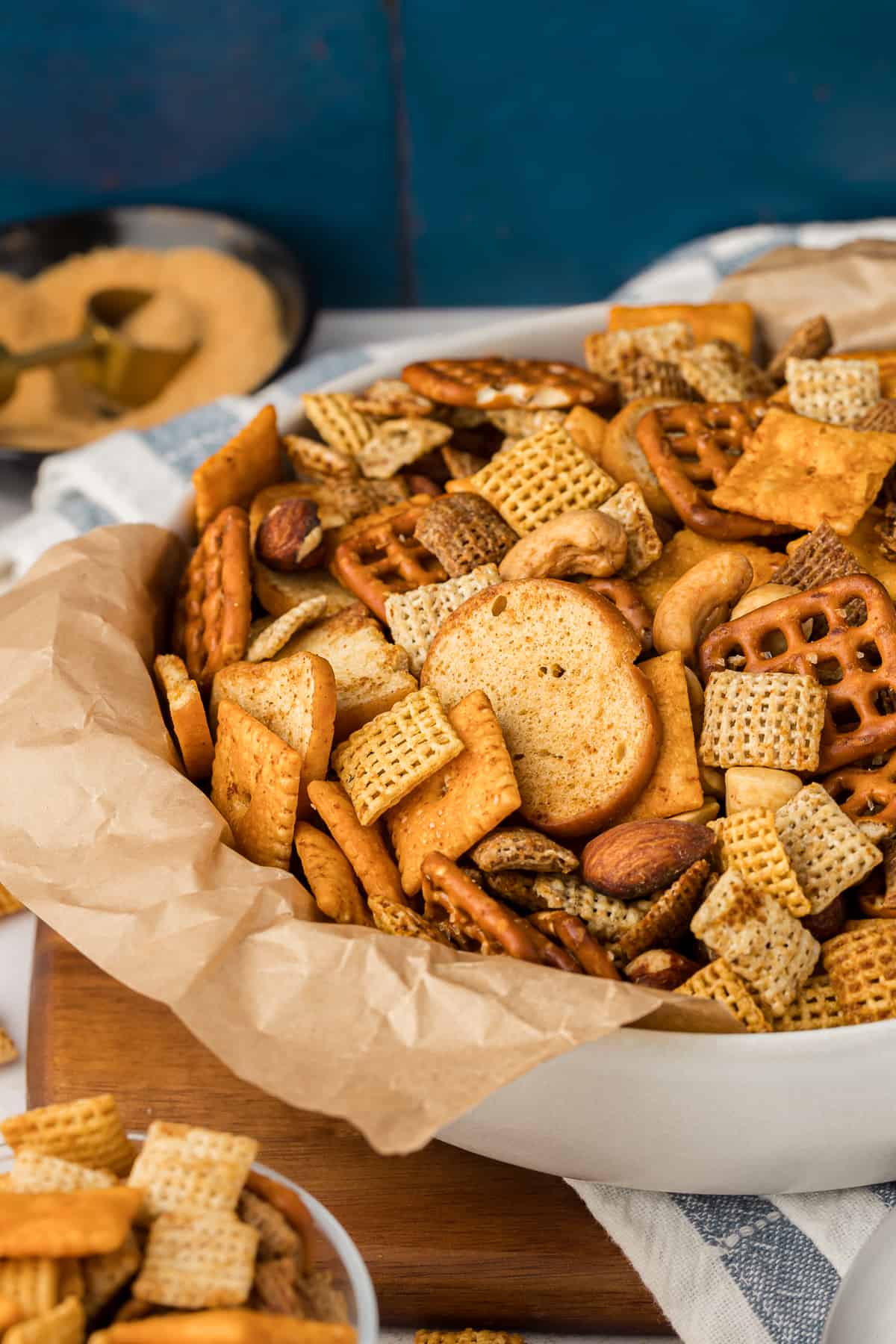 a white bowl lined with brown paper filled with chex mix sitting on a wood piece with a white and blue striped towel on it, a small bowl of chex mix beside it and a small bowel of seasoning in the background