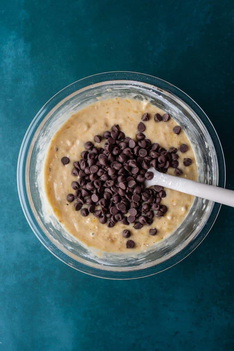 A glass bowl filled with banana bread batter for banana chocolate chip muffins, topped with chocolate chips and a white spatula resting inside, set on a teal surface.
