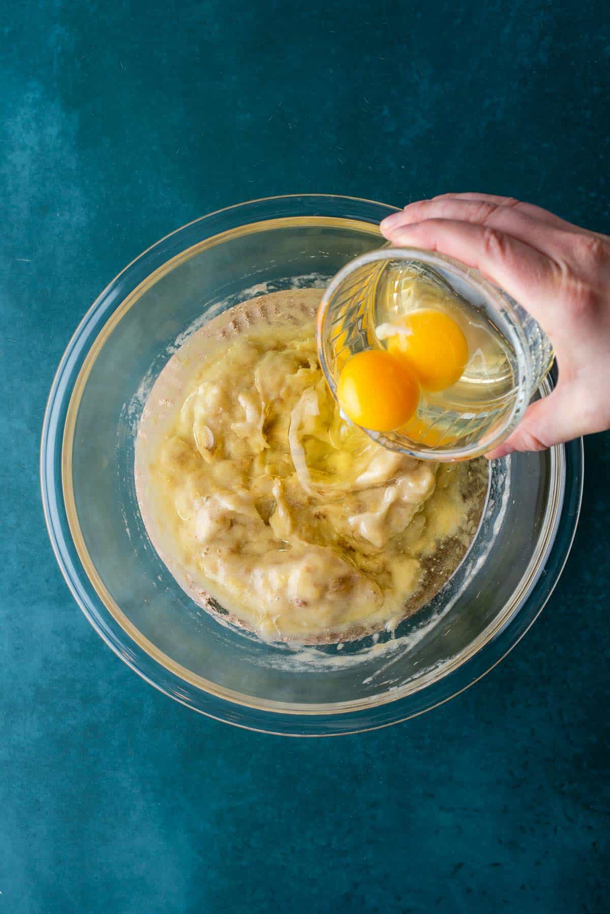 A hand holds a glass bowl with two raw eggs over a larger bowl containing mashed bananas and other ingredients, ready to be combined for banana chocolate chip muffins, on a blue surface.