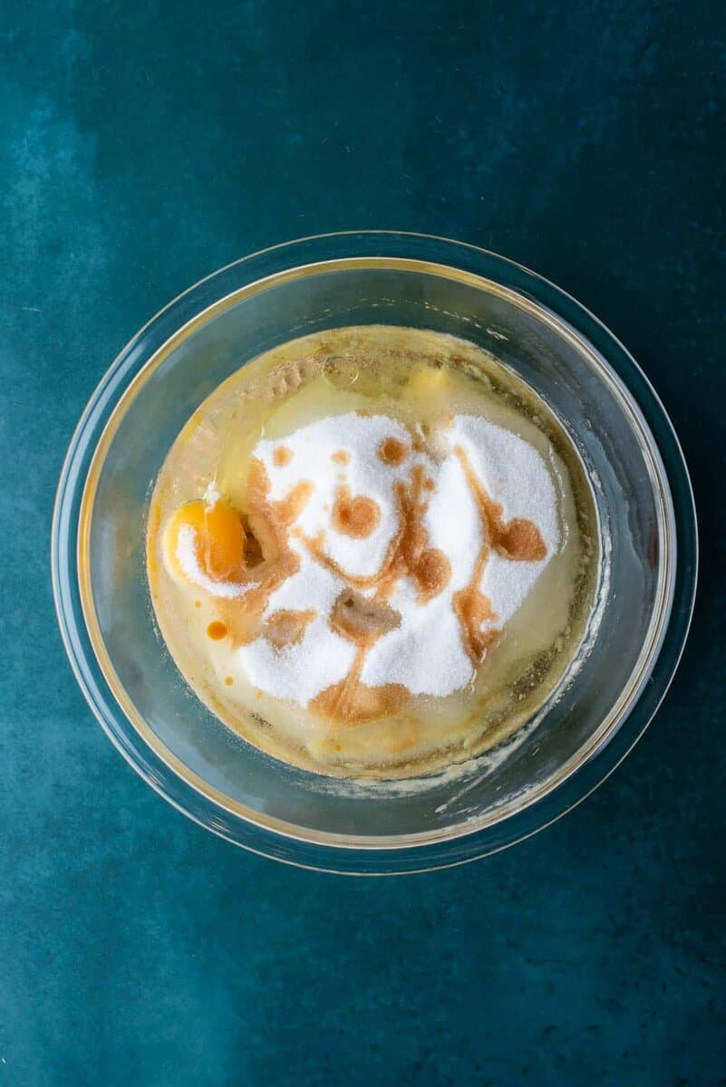 A glass bowl filled with eggs, sugar, and vanilla extract, ready to be mixed for banana chocolate chip muffins, sits on a teal surface.