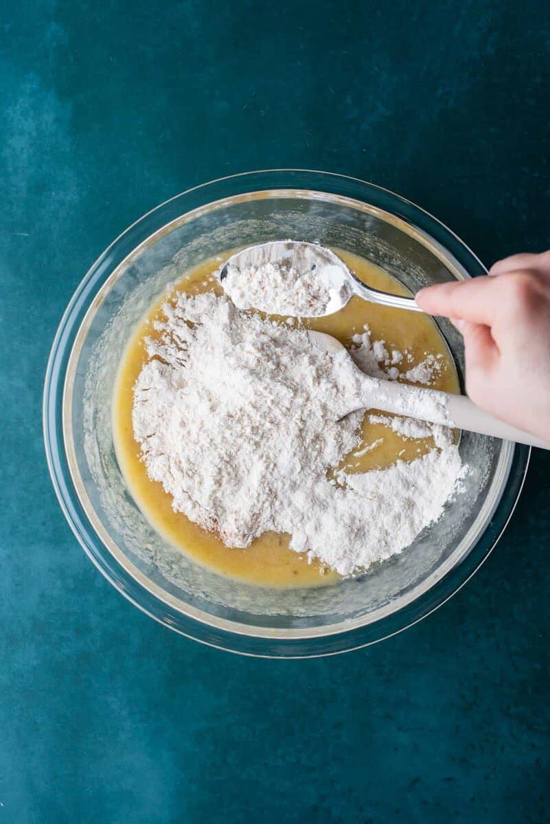 A hand adds flour from a spoon into a glass bowl filled with yellow banana chocolate chip muffin batter, set on a teal surface.