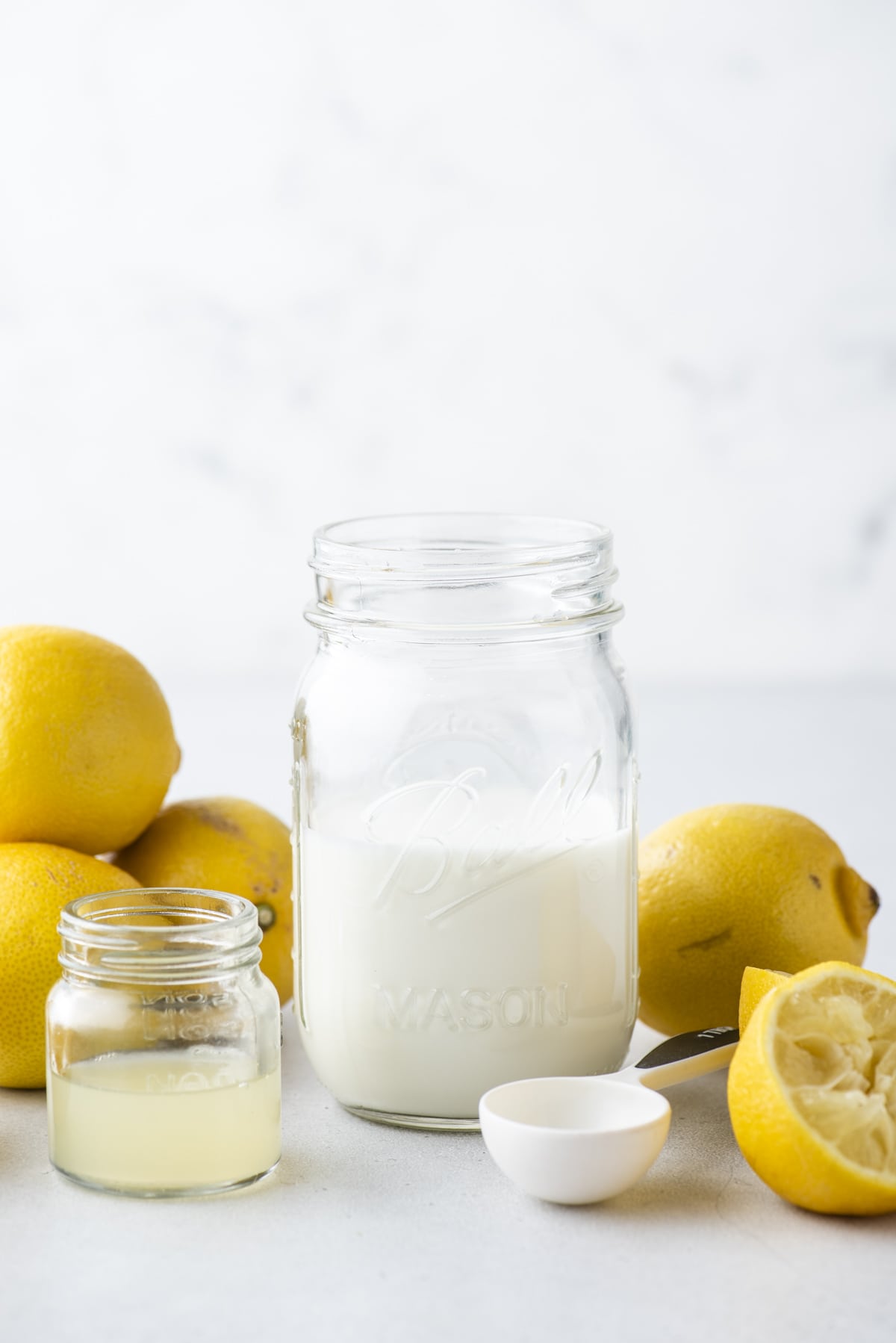 a mason jar full of buttermilk surrounded by fresh whole and sliced lemons, a white tablespoon measurer and a small clear glass jar of lemon juice